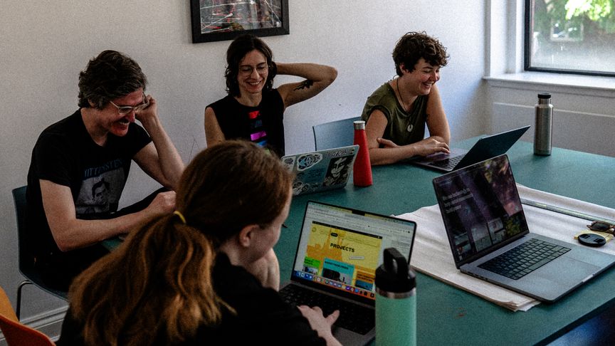 The team working from Pelican House. Four people sit around a table, looking at their laptops and smiling.